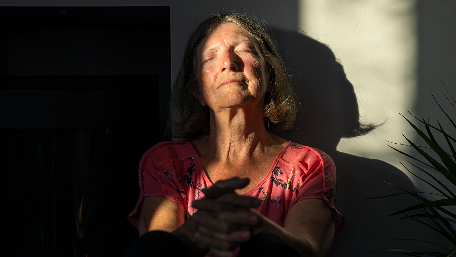 A content senior woman sits against the wall with the last of the day's sun rays warming her face, as she takes time to feel her emotions.