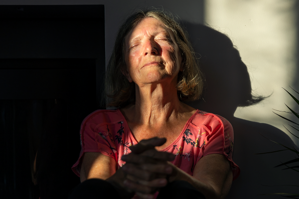 A content senior woman sits against the wall with the last of the day's sun rays warming her face, as she takes time to feel her emotions.