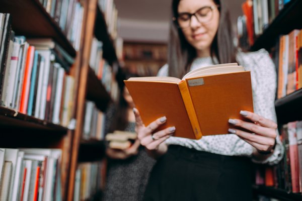 Woman in library standing by bookshelf reading a book | Magellan Healthcare